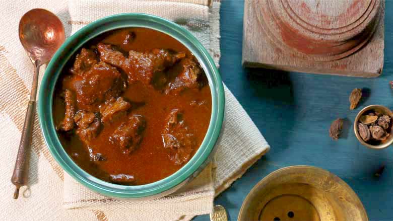 A top view of Rampuri Mutton Korma seen in a emerald green ceramic serving bowl on a cream and gold woven table mat. Lying on a blue wooden table with a few small copper bowls - Taar Gosht Recipe